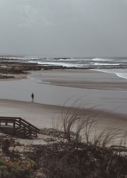 A lone person stands on a wide, calm beach with muted tones, evoking solitude.