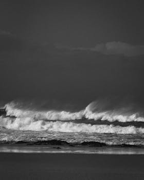 Dramatic black and white seascape with waves crashing on a rocky beach under a moody sky.