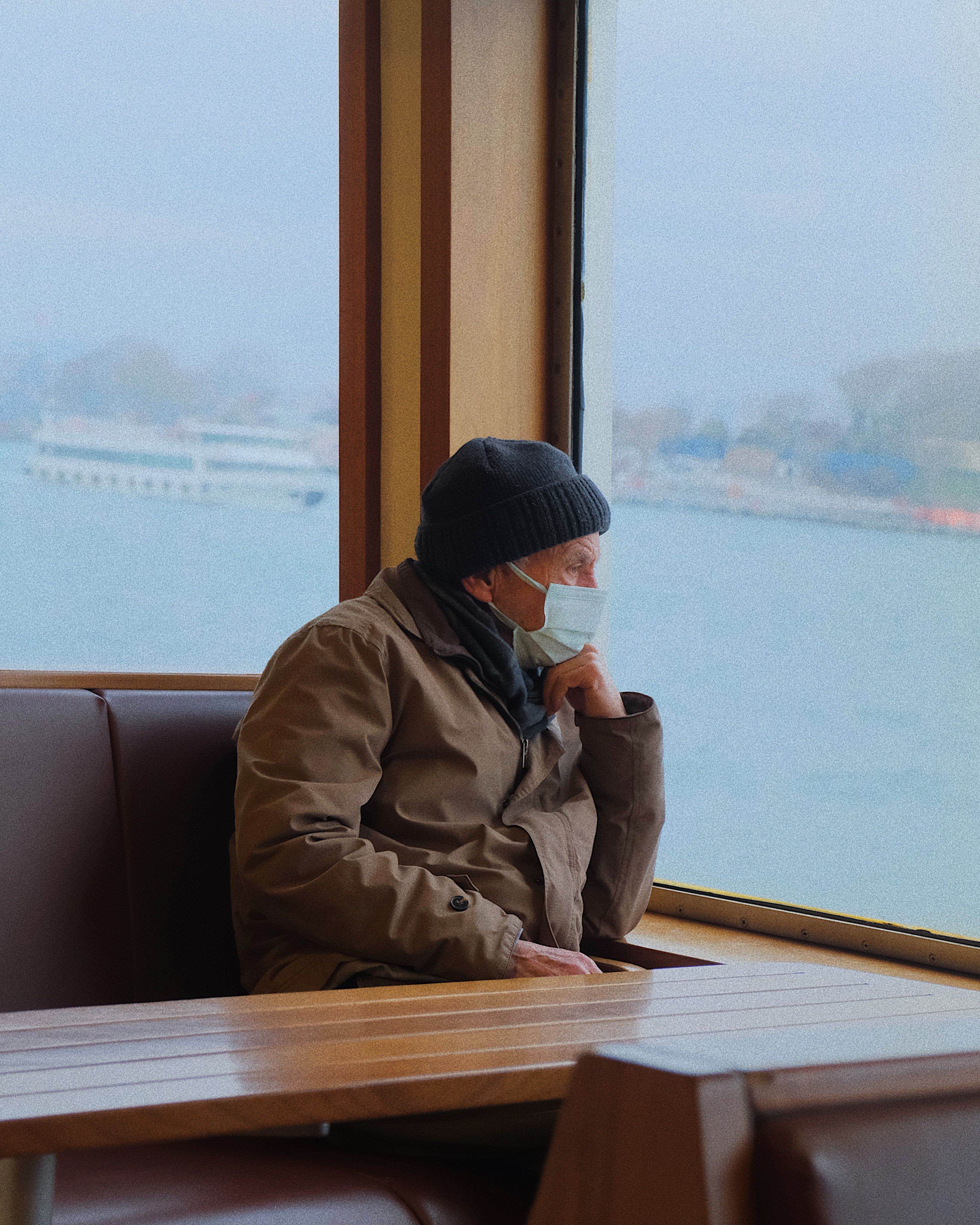 Senior man in a beanie gazes out from an Istanbul ferry window, wearing a face mask.
