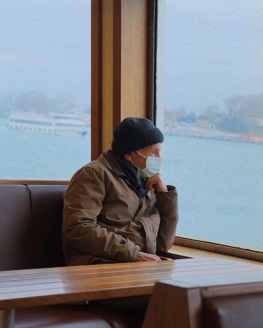 Senior man in a beanie gazes out from an Istanbul ferry window, wearing a face mask.