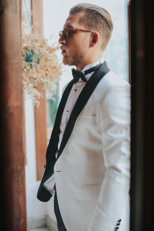 Free Stylish groom wearing a white tuxedo and sunglasses indoors, exuding modern sophistication. Stock Photo