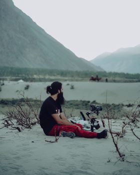 Man sitting in Leh's desert with camera gear, surrounded by scenic mountains.