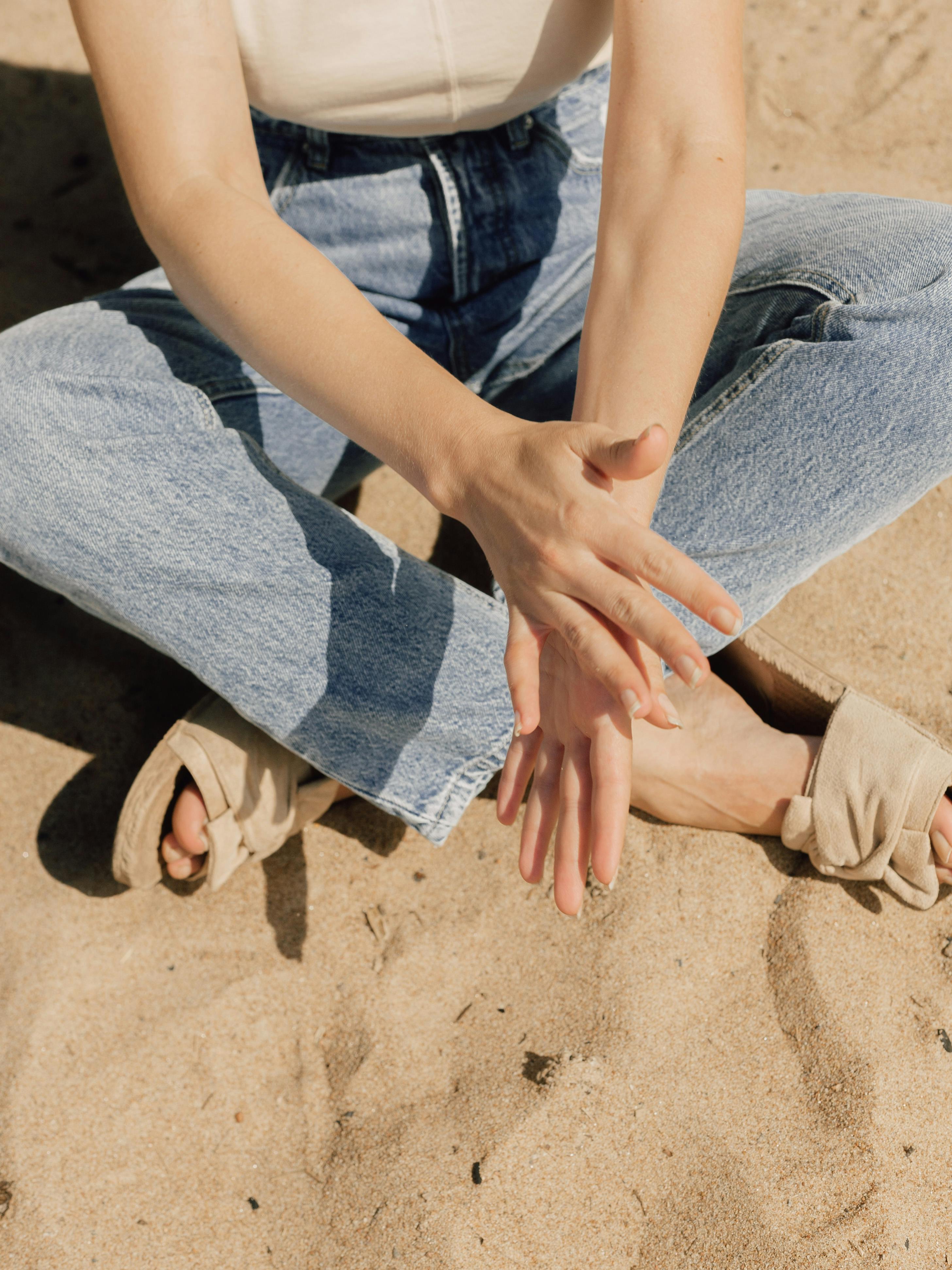 Close-up of a woman sitting on the sandy beach wearing jeans.