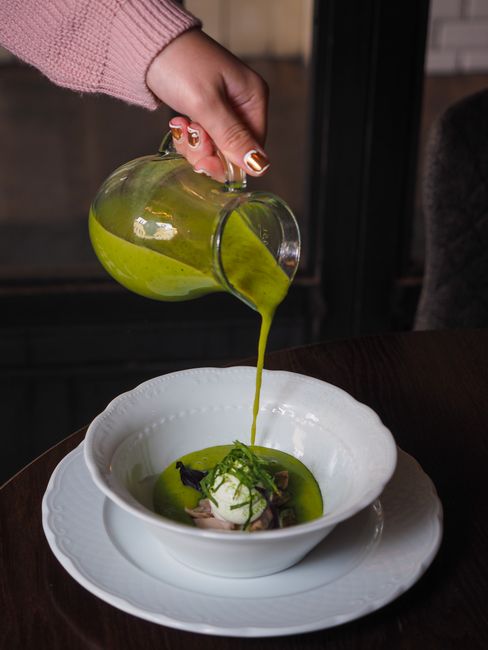 Chef's hand gracefully pours vibrant green sauce over a plated dish in a white bowl, capturing a culinary moment.