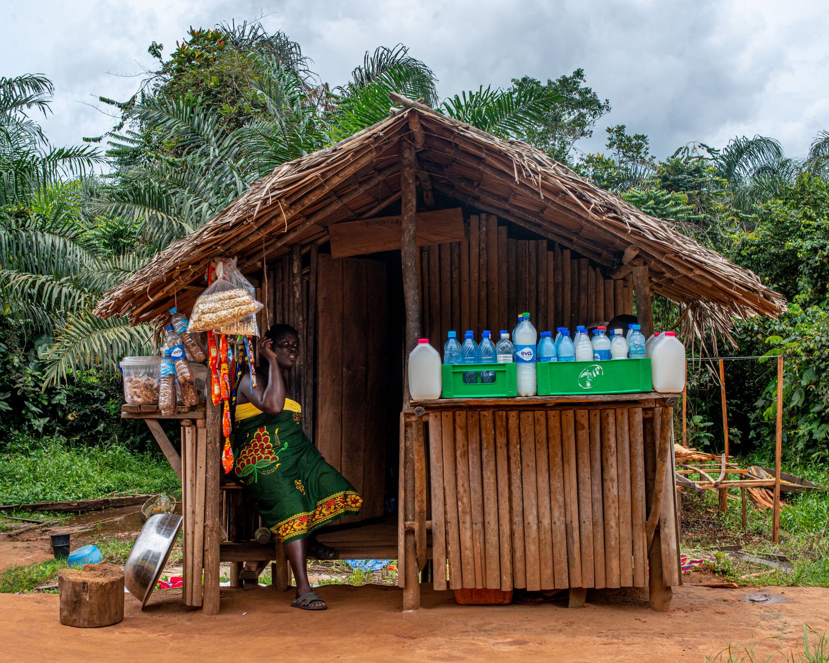 A Nigerian woman in a traditional hut selling bottled refreshments in a rural setting.