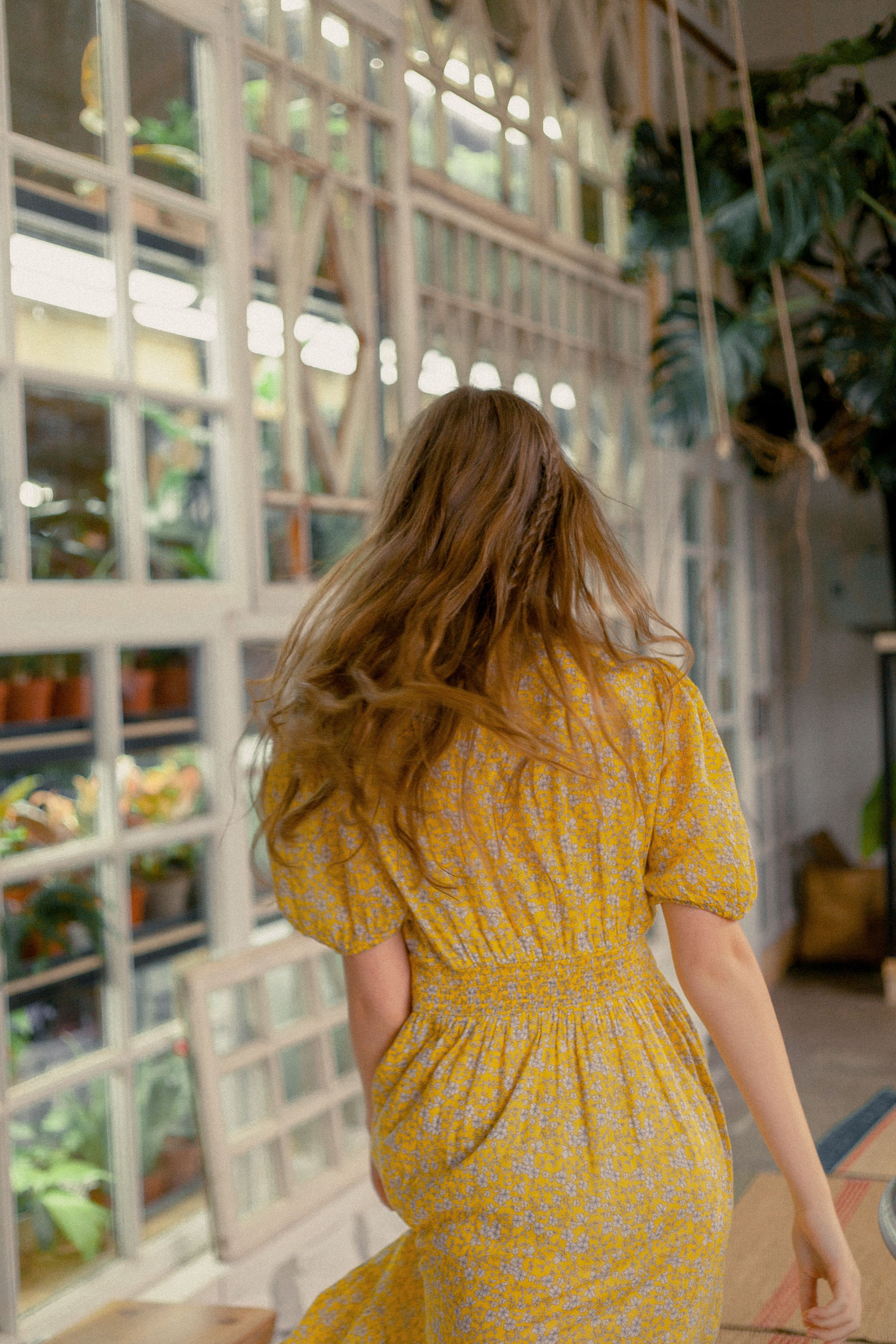 A woman with long brown hair in a yellow floral dress walking indoors, surrounded by plants and windows.