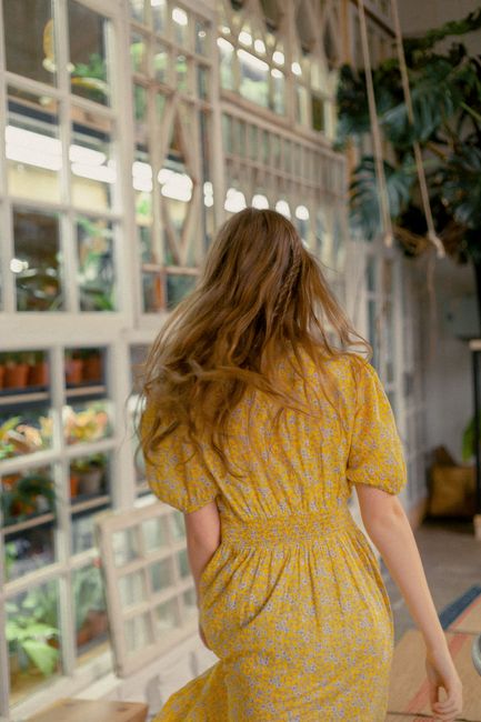A woman with long brown hair in a yellow floral dress walking indoors, surrounded by plants and windows.
