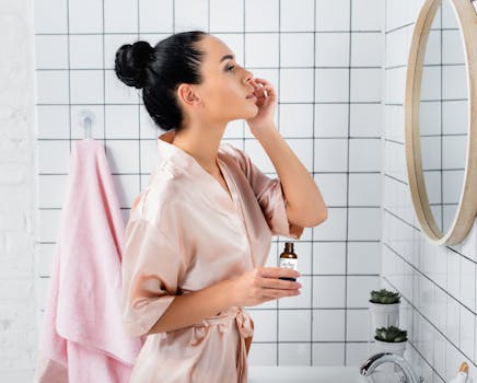 A woman in a bathrobe applying skincare serum in a stylish white tiled bathroom.