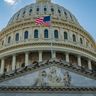 A detailed close-up of the United States Capitol dome and pediment with the American flag flying prominently.