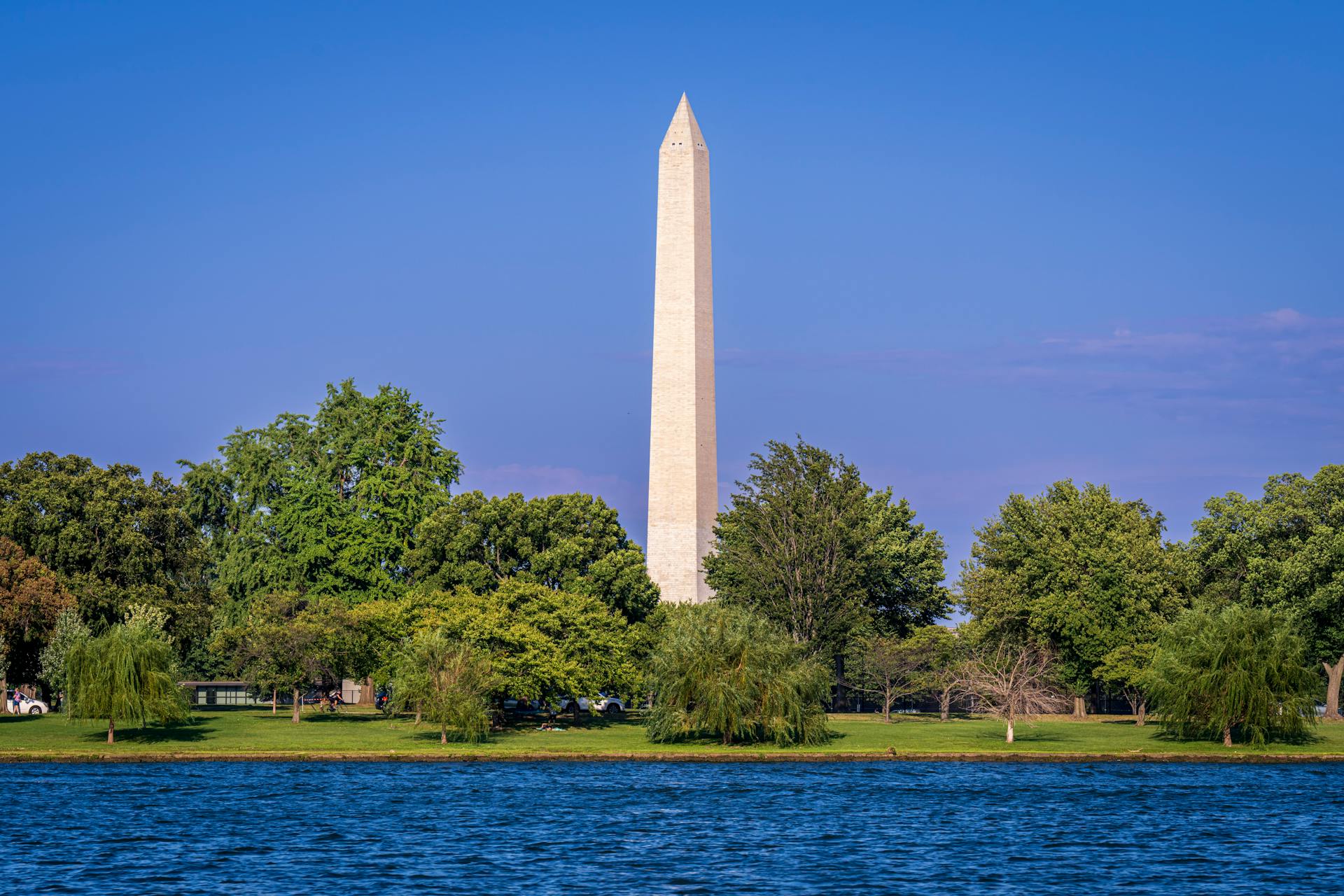 Beautiful view of the Washington Monument surrounded by trees with a clear blue sky and waterscape in Washington D.C.