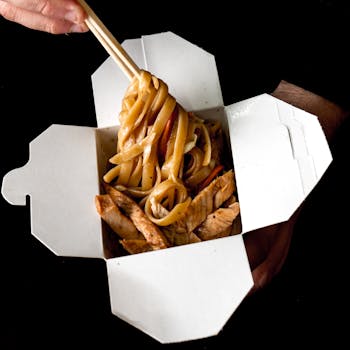 Close-up of hands holding a takeout box of spicy Asian noodles with chopsticks.