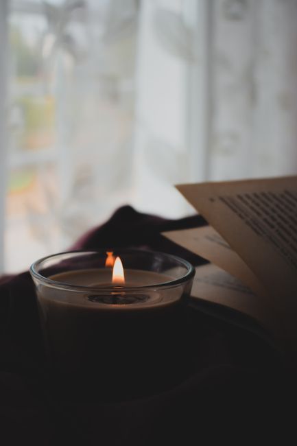 A warm candle flame flickers beside an open book near a window, creating a cozy atmosphere.