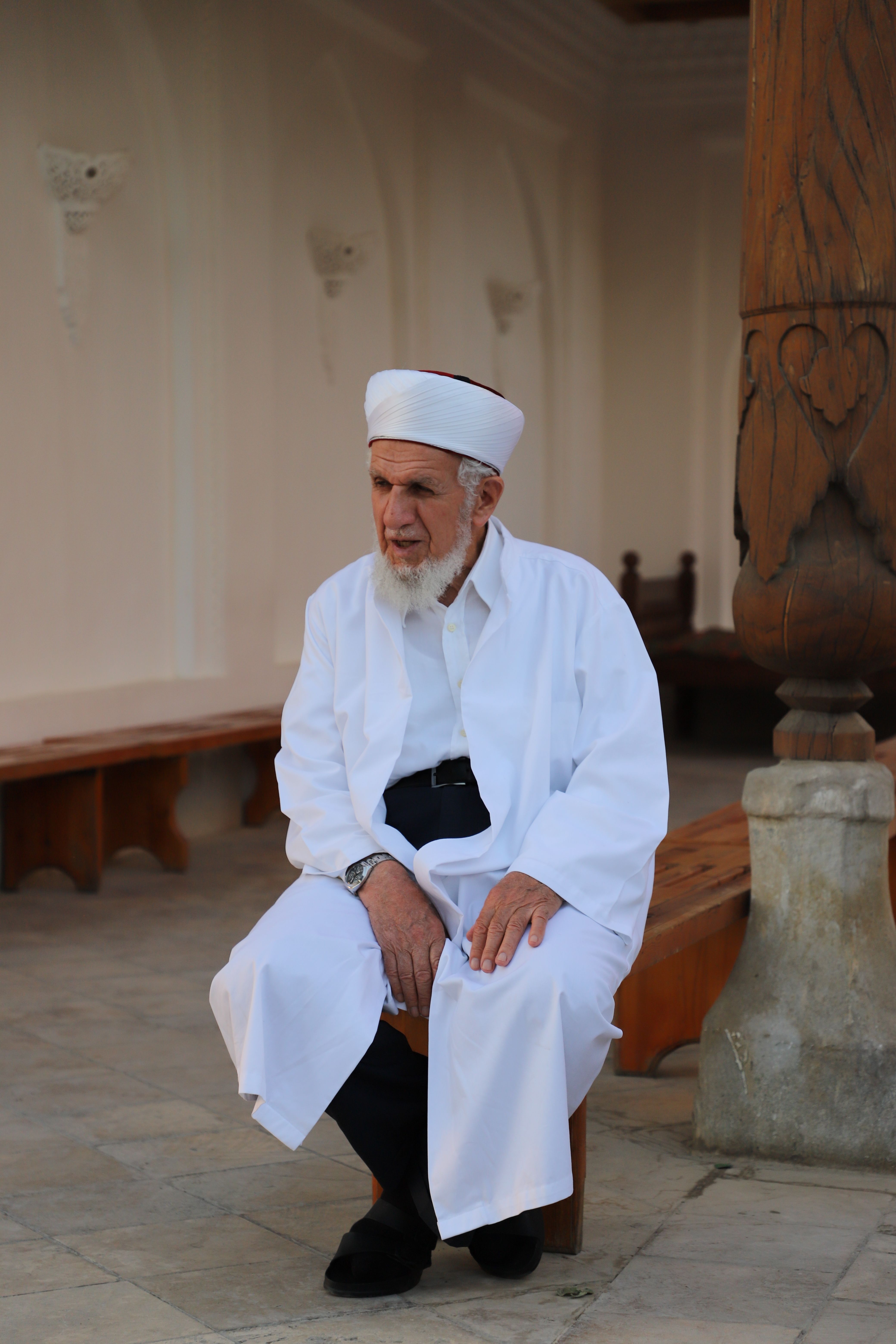 Elderly man in traditional white attire sitting on a wooden bench outdoors, embodying spirituality.
