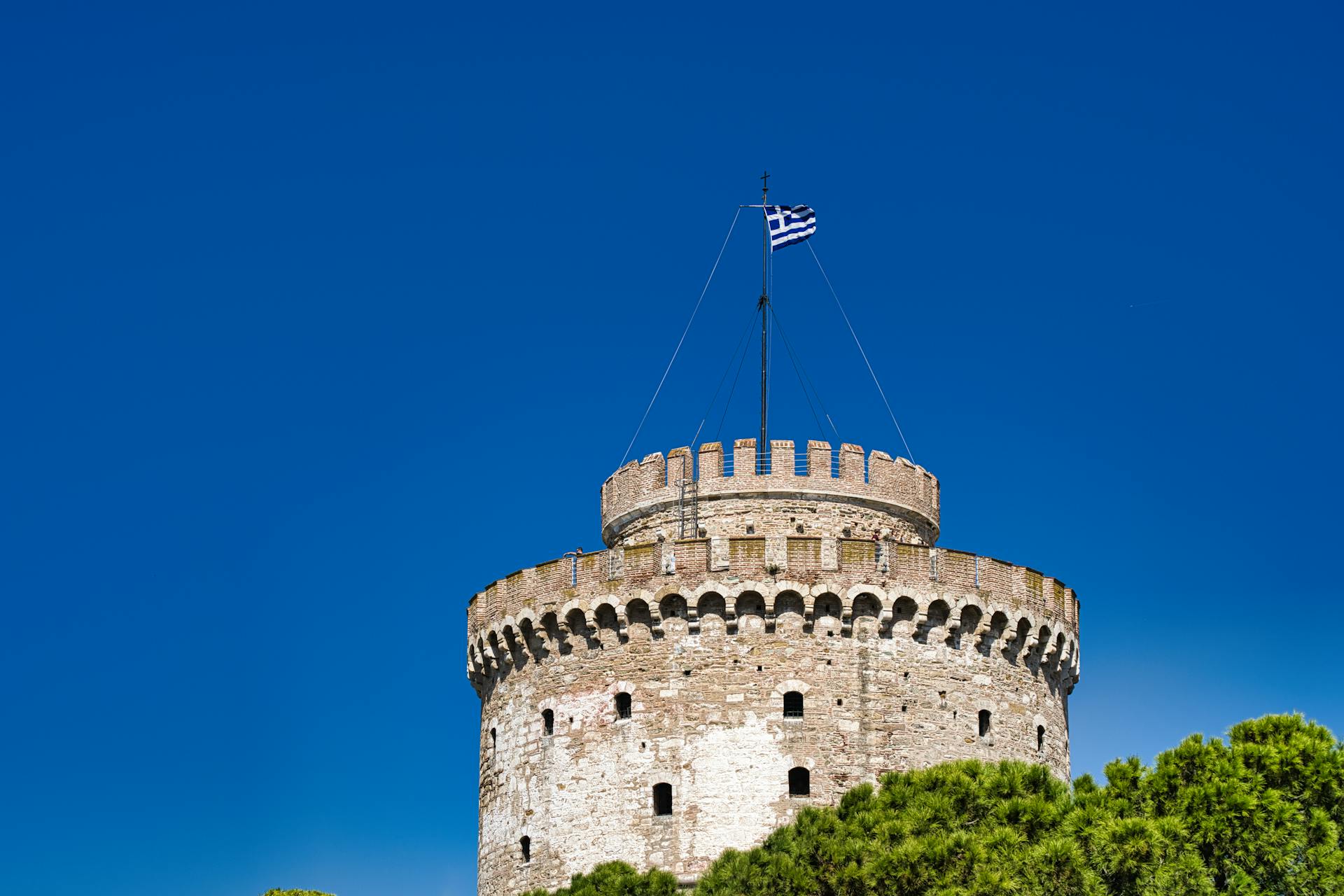 Majestic view of the historic White Tower of Thessaloniki against a bright blue sky with a Greek flag.