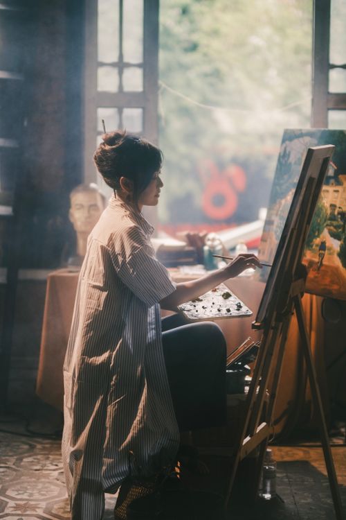 Free Asian woman painting at an easel in a soft-lit art studio, displaying creativity and focus. Stock Photo