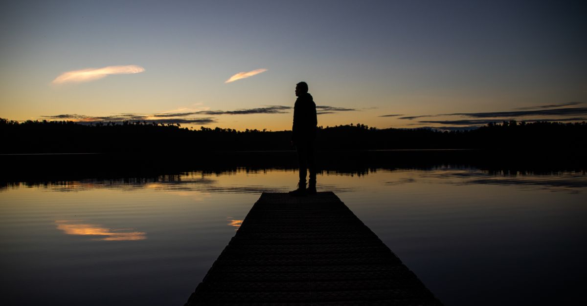 A serene silhouette of a person standing on a dock during sunset, capturing the tranquil reflections in the water.