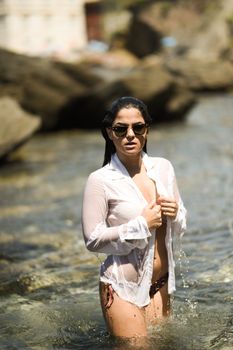Woman standing in clear water, wearing sunglasses and white shirt, enjoying a summer day.