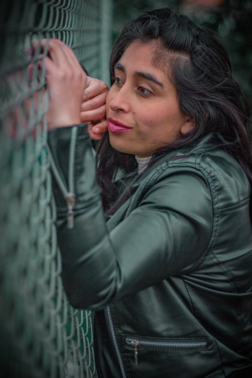 Free Portrait of a thoughtful woman in a leather jacket leaning on a chain link fence outdoors. Stock Photo