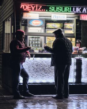 Two men engage in conversation at a street food stall with colorful neon lights at night.