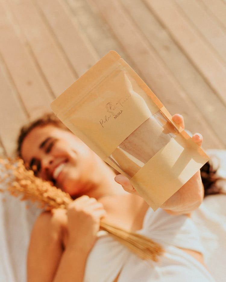 Smiling Woman Lying On A Wooden Floor Holding A Dried Flowers And Bath Salt Refill Pack