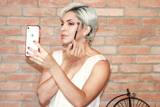 Caucasian woman applying makeup with a brush while using her smartphone as a mirror against a brick wall.