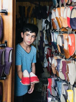 A boy showcases red traditional shoes at a vibrant outdoor market stall.