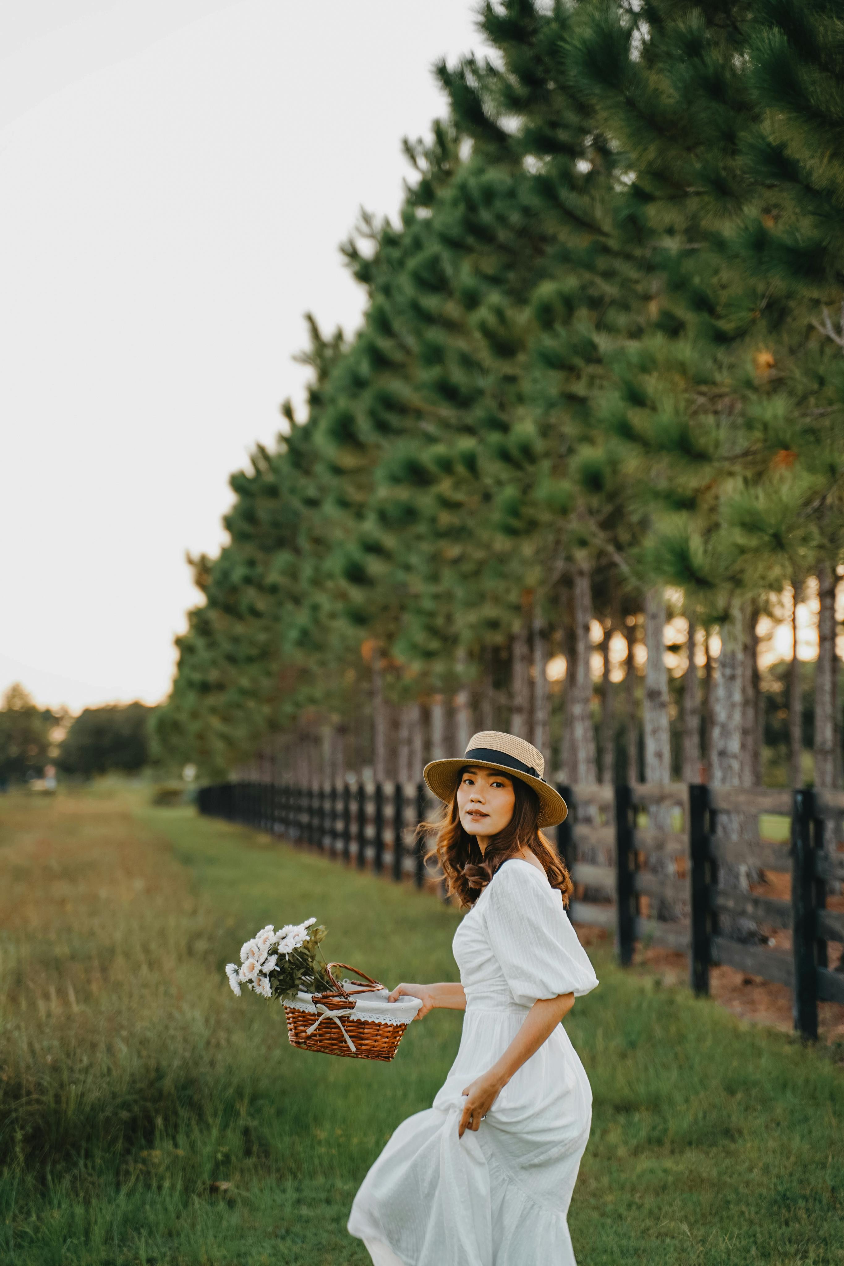 A woman in a white dress and hat walks through a serene field carrying a basket of flowers.