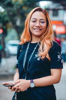 Portrait of a smiling woman with eyeglasses holding a smartphone on a bustling street in Dhaka.
