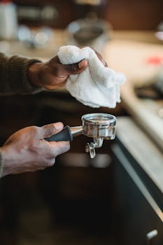 Close-up of a barista's hands cleaning a portafilter with a cloth in a coffee shop setting.