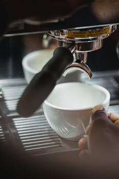 Hand operating a coffee machine pouring espresso into a white cup.