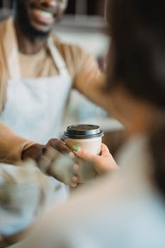 Barista in apron hands coffee to customer in cozy cafe setting, warm interaction.