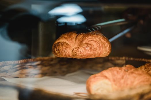 A fresh croissant is lifted from a bakery display with tongs, showcasing golden brown layers.