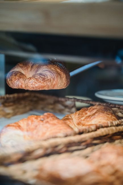 A row of golden, flaky croissants with visibly laminated layers on a wooden bakery board, one torn open to show the honeycomb interior.