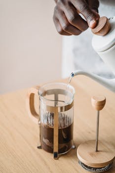 An adult's hands pouring hot water into a French press on a wooden table.