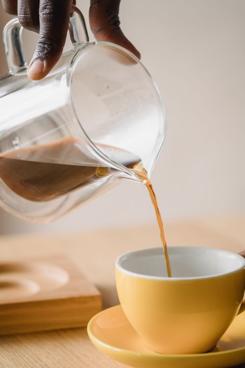 Free Hand pouring coffee from glass pitcher into a yellow cup on a wooden table. Stock Photo
