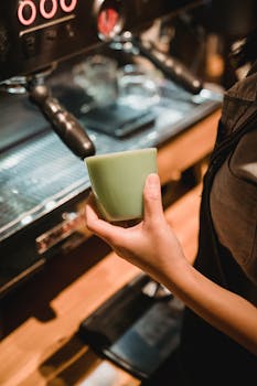 Barista holding a green cup near an espresso machine in a cozy café setting.