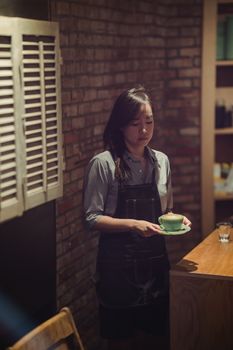 Female barista standing with coffee cup in a cozy, dimly lit cafe setting.