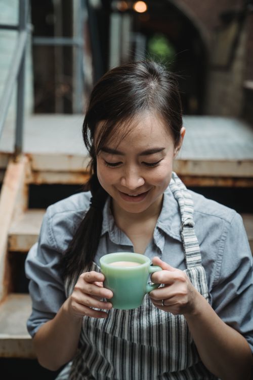 Free A woman in an apron smiles while holding a mug of coffee, sitting on steps. Stock Photo