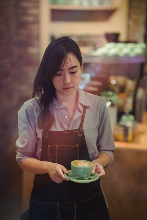Free Young female barista holding a latte art coffee cup in a warm and inviting café environment. Stock Photo