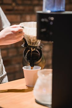 Close-up of a barista pouring freshly brewed coffee into a paper cup in a cafe setting.