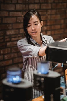 Female barista in striped apron preparing coffee with machines in warm cafe setting.