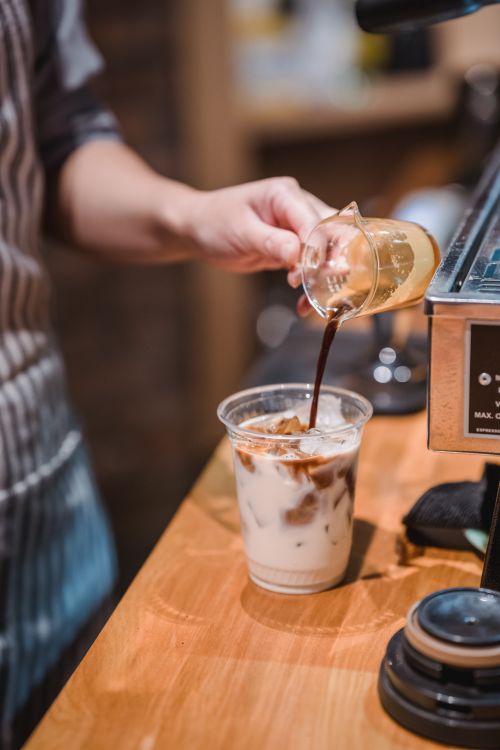 Free Barista pouring coffee into a plastic cup with ice and milk on a wooden counter. Stock Photo