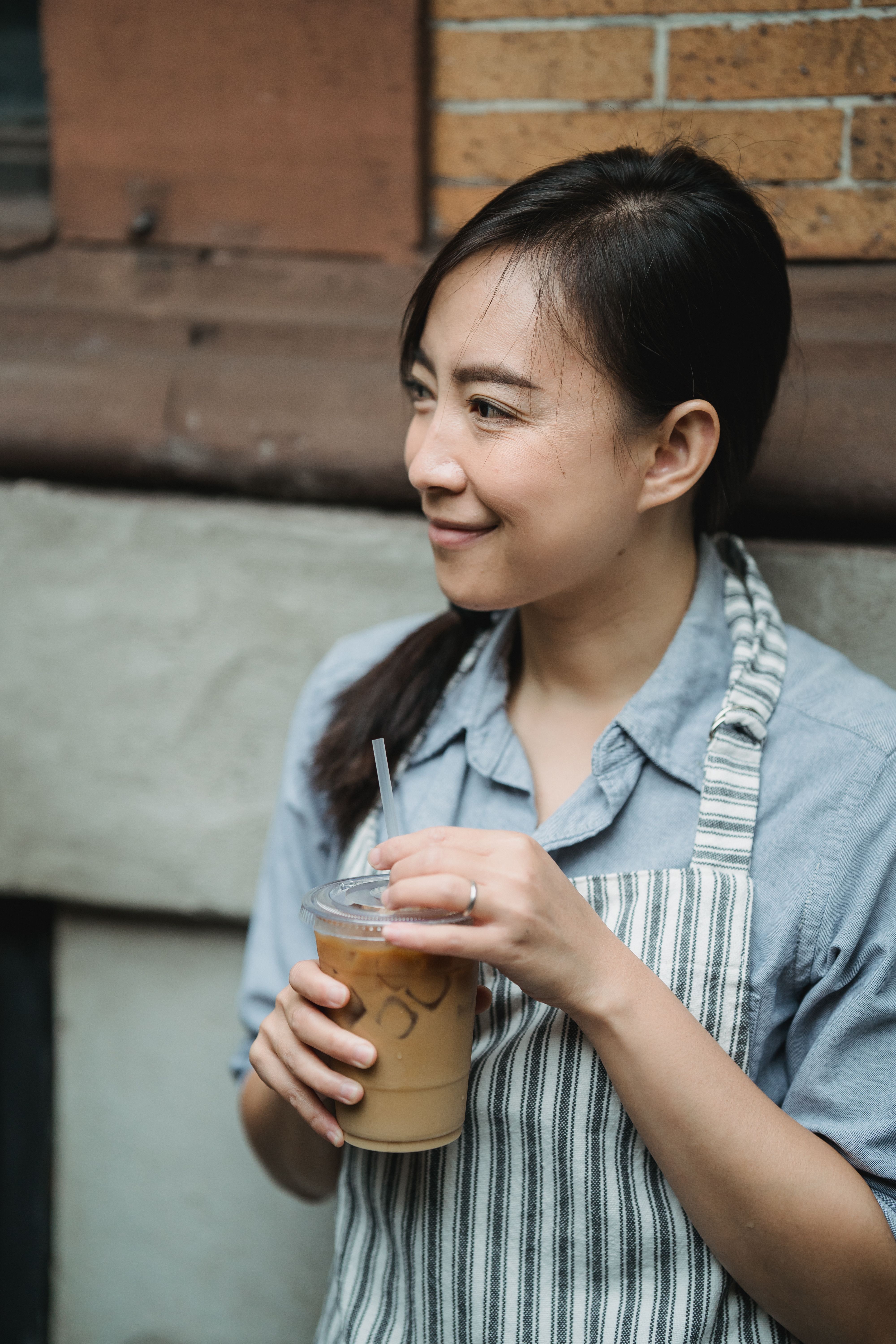 Smiling woman in striped apron holding iced coffee outside on a nice day.