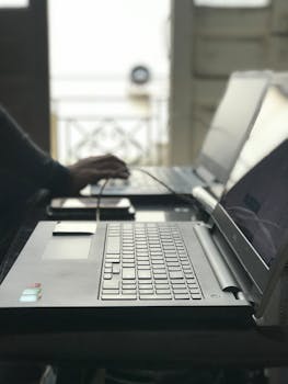 A backlit image of a person typing on laptops at a table, showcasing technology in use.
