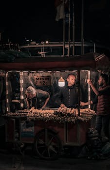 Three men at a bustling street food stall selling corn at night. Atmospheric lighting enhances the market scene.