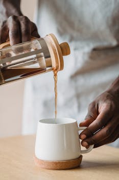 Barista pouring piping hot coffee from a french press into a white mug indoors.