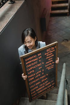 Smiling barista carrying a chalkboard menu on stairs inside a cafe, portraying a lively atmosphere.