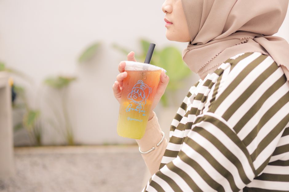 Side view of a woman in a hijab sipping an iced tea outdoors, wearing a striped shirt.