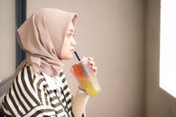 Side profile of a Muslim woman in a hijab drinking a cold beverage indoors. Neutral tones and soft lighting.