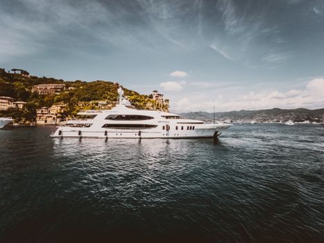 Stunning luxury yacht cruising by picturesque waterfront town, nestled by verdant hills under a clear sky.